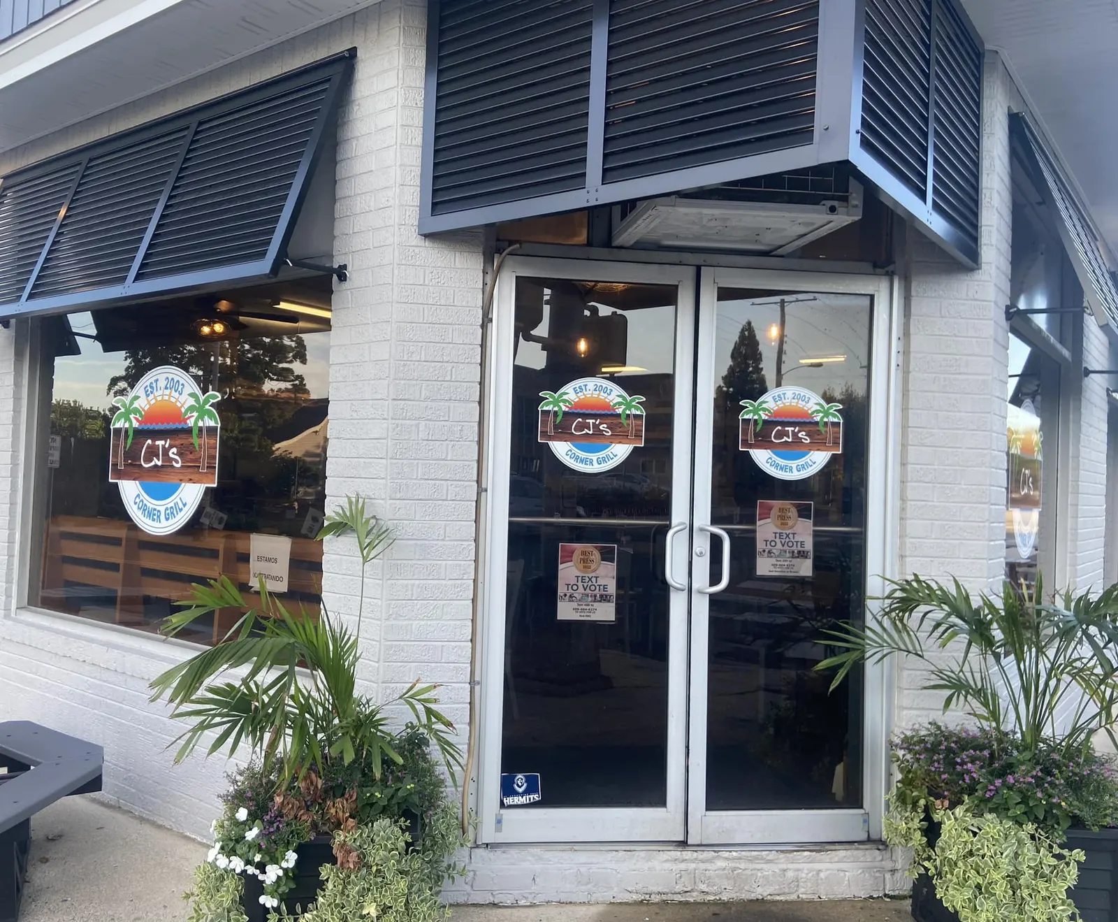 CJ's Corner Grill entrance with blue awnings, wooden seating and potted palms